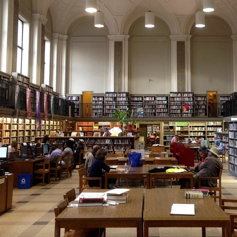 Tall cases of books surround a central area full of desks as people sit reading and accessing information from computers in a public library.